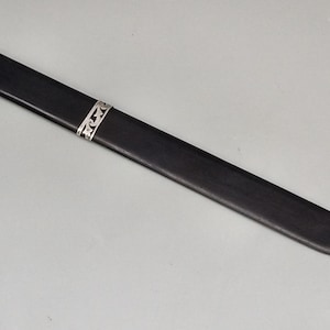 Puede incluir: Una funda de cuchillo de madera negra con una banda decorativa plateada. La funda es larga y delgada, con un extremo puntiagudo. La banda plateada presenta un patrón intrincado y repetitivo. El artículo se muestra sobre un fondo gris claro liso.