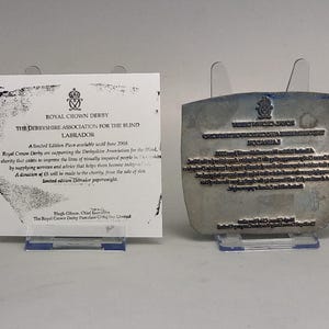 May include: A white rectangular paper certificate from Royal Crown Derby regarding a limited edition Labrador paperweight, displayed next to a metallic printing plate featuring the same text in reverse, both held upright by clear plastic stands.