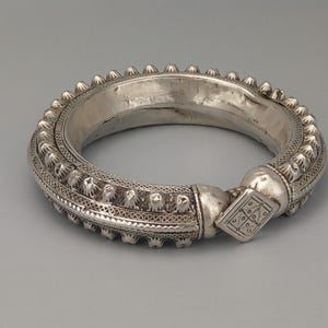 May include: An intricately designed silver bangle. The thick band is adorned with detailed patterns and small, pointed studs along the outer edge. A square clasp, decorated with a geometric design, fastens the bracelet.