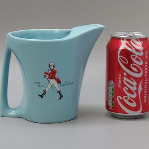 May include: A light blue ceramic pitcher with a handle and a spout. The pitcher features a graphic of a man in a red coat and the text "BORN 1820 - STILL GOING STRONG". A red Coca-Cola can is next to the pitcher.