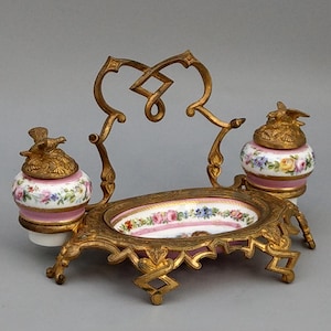 Könnte beinhalten: Ein antikes Tintenfass-Set mit einem goldfarbenen Metallrahmen und floralen Porzellan-Tintenfässern. Die Tintenfässer haben vogelartige Deckel und sind mit rosa, blauen und gelben Blumen verziert. Das Set enthält eine ovale Schale mit floralem Design.