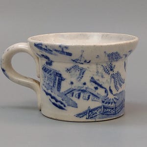 Op de afbeelding: Een kleine, antieke witte keramische mok met een blauw Willow-patroon. De mok heeft een gebogen oor en een licht uitlopende rand. Het blauwe patroon toont een landschap met gebouwen en bruggen.