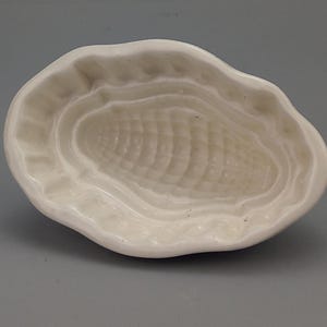 Puede incluir: Un molde de cerámica color blanco hueso que presenta un diseño texturizado y hundido de una mazorca de maíz en el centro. El molde tiene un borde exterior irregular, ondulado y redondeado con un acabado liso y brillante en todo.