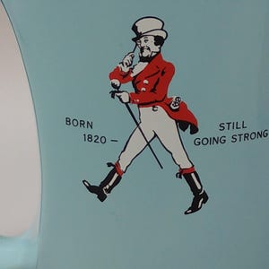May include: A light blue mug featuring a graphic of a man in a red coat and white pants, holding a cane. Text on the mug reads "BORN 1820 - STILL GOING STRONG". The mug has a curved handle.