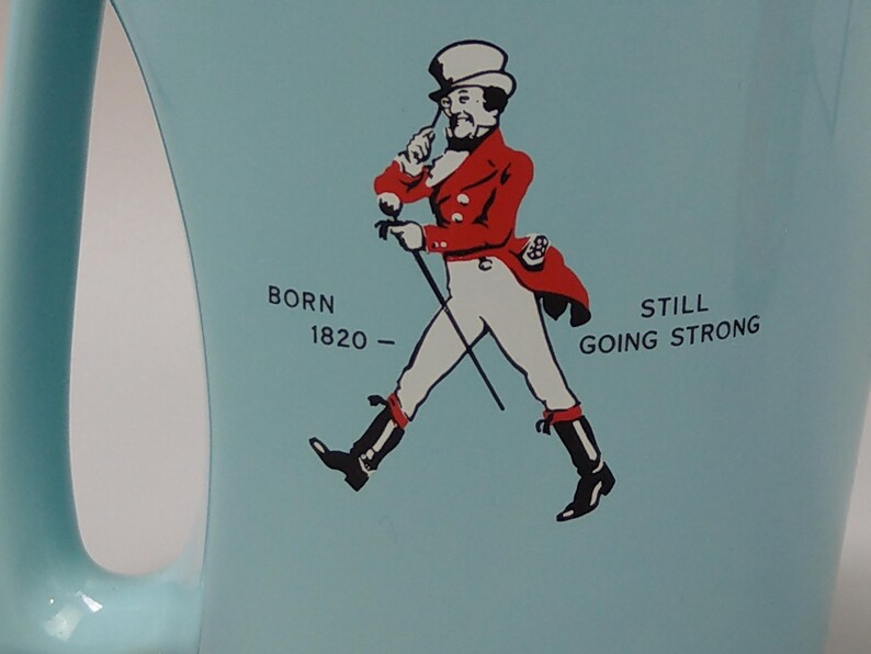 May include: A light blue mug featuring a graphic of a man in a red coat and white pants, holding a cane. Text on the mug reads "BORN 1820 - STILL GOING STRONG". The mug has a curved handle.