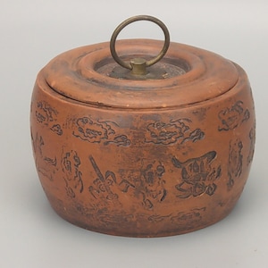 May include: A terracotta jar with a lid and a brass handle. The jar is decorated with a brown glaze and has a design of clouds and figures.