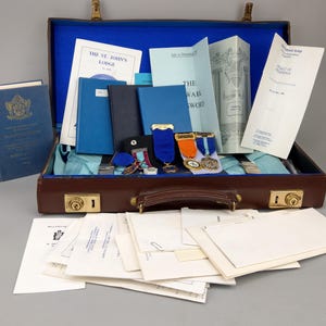 May include: A brown briefcase with gold-tone latches containing blue-lined interior, multiple blue books, pamphlets titled The St. John's Lodge and The Mark Sword, and several medals with ribbons. A stack of white paper documents lies in front of the case.