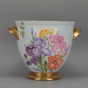 Puede incluir: Un recipiente de porcelana blanca con un colorido arreglo floral de lirios morados, un lirio rosa y flores naranjas. El contenedor tiene dos asas laterales en tono dorado y una base circular dorada a juego con borde.