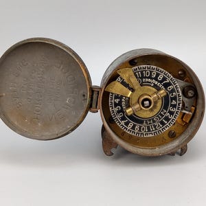 May include: An open, antique metal clock with a hinged lid. The clock face features a black and gold dial with numbers and the word "NIGHT". The lid has etched text. The clock is aged with a weathered appearance.