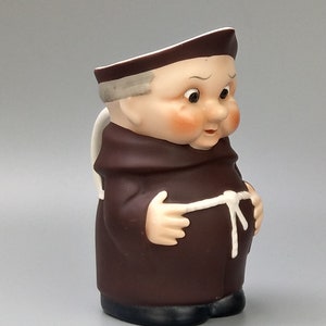 May include: A brown ceramic pitcher shaped like a monk with a white rope belt and a white face with brown eyes.