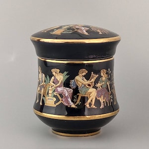 Può includere: Un vaso in ceramica nera con bordo dorato e coperchio. Il vaso è decorato con una scena di figure greche che suonano musica.