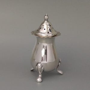 May include: A silver-colored, antique-style salt shaker with a decorative lid featuring small holes. The shaker has a bulbous body and rests on three small, curved feet. The lid is topped with a small, ornate finial.