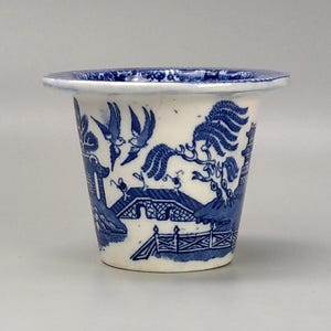 May include: A small, white ceramic planter with a blue and white willow pattern. The planter has a wide, flat rim and a rounded base.
