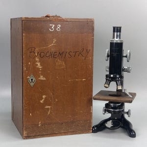 Könnte beinhalten: Ein schwarzes Metallmikroskop mit silbernem Okular und mehreren Objektivlinsen, das neben einem rechteckigen Aufbewahrungskoffer aus Holz steht. Der Koffer trägt die handschriftliche Aufschrift "38" und "BIOCHEMISTRY" sowie ein Schlüsselloch.