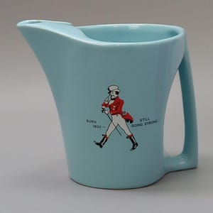 May include: Light blue ceramic pitcher with a handle and a pouring spout. The pitcher features a graphic of a man in a red coat and the text "Born 1820 - Still Going Strong."