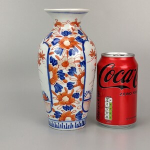 May include: A small, white porcelain vase with a blue and orange floral pattern. The vase is approximately 15 cm tall and has a narrow neck. It is sitting on a white surface next to a can of Coca-Cola.