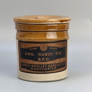 Puede incluir: Un tarro de cerámica vintage con tapa. El tarro es de color marrón claro y crema con una etiqueta oscura que dice "UNG. RUSCI CO. B.P.C." y "JAMES WOOLEY SONS & CO LTD MANCHESTER". El tarro es probablemente para almacenamiento.
