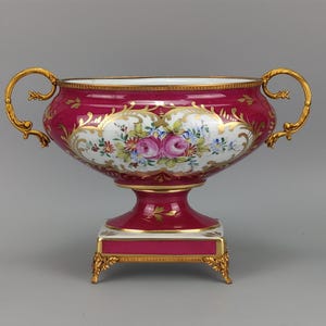 Puede incluir: Un cuenco de porcelana magenta con dos asas doradas ornamentadas y una base cuadrada. El cuenco presenta un cartucho blanco central decorado con un ramo de flores pintado a mano de rosas rosas y flores azules, con detalles en pan de oro.