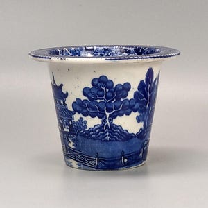 May include: A small, white ceramic planter with a blue and white floral design. The planter has a wide rim and a slightly flared base.