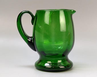 May include: A green glass pitcher with a rounded body, a curved handle, and a pouring spout. The pitcher has a wide base and a smooth, glossy surface. The pitcher is approximately 6 inches tall.