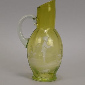 May include: A vintage, pale yellow glass jug with a clear handle. The jug features an etched design of a person and a landscape. The top of the jug is cut at an angle.