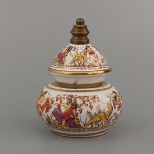 Può includere: Un vaso di ceramica bianca con un coperchio dorato e un bordo dorato. Il vaso è decorato con una scena di persone e animali in un ambiente boschivo.