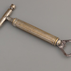 Pode incluir: Um abridor de garrafas vintage com um design semelhante a um martelo. O abridor apresenta um corpo prateado com uma seção central texturizada dourada e uma extremidade pontiaguda. A extremidade oposta tem um abridor de garrafas.