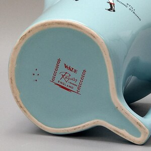 May include: A light blue Wade Regicor pitcher with a handle and a spout. The bottom of the pitcher features the Wade Regicor London England logo in red. The pitcher has a white rim and the words "STILL GOING STRONG" are visible.