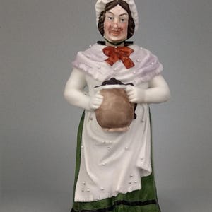 May include: A ceramic figurine of a woman in a white apron and green skirt, holding a brown jug. She wears a white bonnet with a red bow, a lavender shawl, and red shoes. The figurine has a detailed, hand-painted appearance.