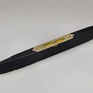 Puede incluir: Un nivel de madera negro vintage con una placa de latón y dos viales amarillos. El nivel mide aproximadamente 30 cm de largo, con forma rectangular y extremos angulados. Es una herramienta de medición.