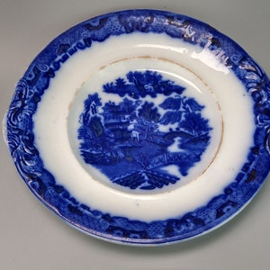 May include: A blue and white porcelain saucer with a decorative floral pattern and a central scene of a house and trees.