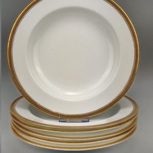 May include: A stack of white ceramic plates with a gold Greek key border. The plates have a shallow bowl shape and are stacked on top of each other. The gold border adds a touch of elegance to the tableware.