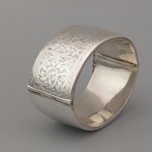 May include: A silver hinged bangle bracelet with an intricate floral design etched into the surface.