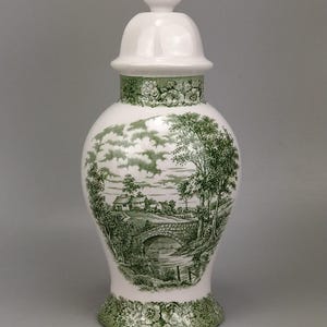 Peut inclure: Un pot à gingembre en céramique blanche avec couvercle assorti, orné d'un motif paysager vert imprimé représentant un pont en pierre, des arbres et des maisons. La base et le col présentent une bordure florale détaillée de couleur verte.