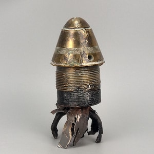 May include: A metallic artillery fuse component featuring a polished brass conical top section with visible horizontal grooves and a small circular aperture. The lower portion consists of a dark, threaded cylindrical base attached to jagged, torn metal fragments.