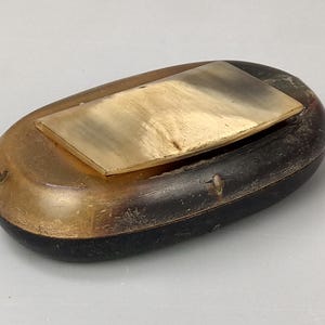 Puede incluir: Un recipiente antiguo, de forma ovalada, con una base marrón oscuro y una parte superior dorada. Una placa rectangular de color dorado está incrustada en la parte superior. El recipiente tiene un aspecto desgastado y envejecido.