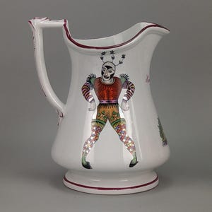 Può includere: Una brocca in ceramica bianca con un'illustrazione dipinta a mano di una figura di giullare mascherato, che indossa un abito rosso, arancione e a quadri multicolori. La brocca presenta un manico curvo e un bordo rosso scuro sulla base e sul bordo.