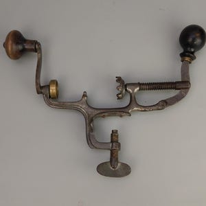 Könnte beinhalten: Ein antiker Obstentkerner aus Metall mit einem Holzgriff und einem schwarzen Knopf. Der Entkerner hat einen Schraubmechanismus und einen Metallring.