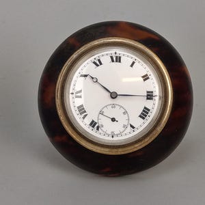 May include: A vintage pocket watch with a white face, black Roman numerals, and a smaller seconds dial. The watch has a gold-colored bezel and is encased in a brown, tortoiseshell-patterned material.