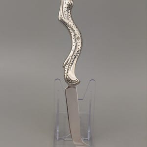 May include: A silver-toned metal knife featuring a curved handle shaped like a stylised leopard with a spotted texture. The blade is smooth, polished, and slightly curved, extending from the base of the leopard-shaped handle to a pointed tip.