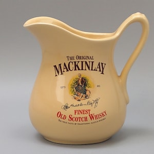 Op de afbeelding: Crèmekleurige keramische kan met een handvat en een schenktuit. De kan heeft de tekst "THE ORIGINAL MACKINLAY FINEST OLD SCOTCH WHISKY" met een illustratie van een man in kilt. Een decoratief item in vintage stijl.