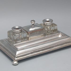 Puede incluir: Un juego de tintero plateado con dos tinteros de cristal y un compartimento con tapa. La base rectangular tiene un borde decorativo y descansa sobre cuatro patas esféricas. Un pequeño adorno plateado remata el compartimento con tapa.