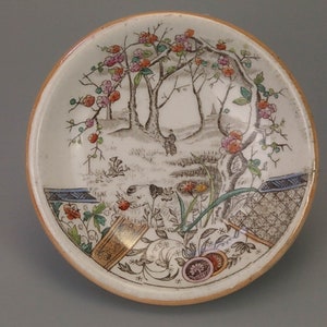 May include: A white ceramic saucer with a floral design. The saucer features a scene of trees with pink flowers and a brown fence. The saucer has a gold rim.