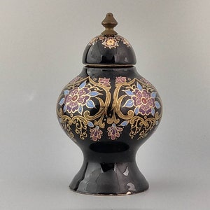 Può includere: Un vaso in ceramica nera con un design floreale dorato. Il vaso ha un coperchio con un pomo dorato.