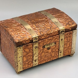 May include: A small, rectangular copper box with a hinged lid. The box is decorated with brass accents and has a raised, textured surface. The lid has a brass plaque with the word "TEA" engraved on it.