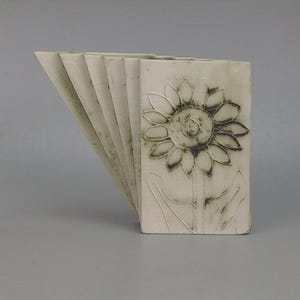 May include: A set of six off-white ceramic rectangular vases arranged in a fan shape. Each vase features a debossed sunflower design with dark grey detailing on the petals and stem, creating a cohesive floral pattern across the collection.
