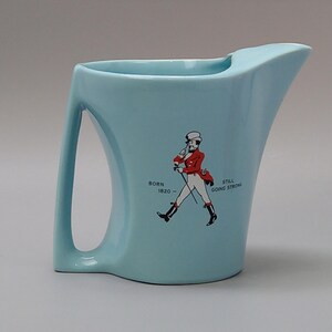 May include: A light blue ceramic pitcher with a curved handle and spout. The pitcher features a graphic of a man in a red coat and the text "BORN 1820 - STILL GOING STRONG."