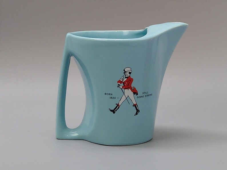 May include: A light blue ceramic pitcher with a curved handle and spout. The pitcher features a graphic of a man in a red coat and the text "BORN 1820 - STILL GOING STRONG."