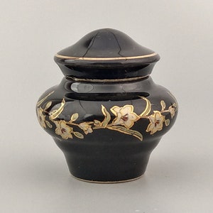 Può includere: Un piccolo vaso in ceramica nera con coperchio, decorato con un motivo floreale dorato e bianco.