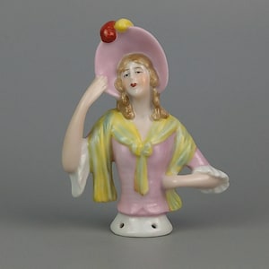 Puede incluir: Una muñeca de porcelana para alfiletero con un vestido rosa, una bufanda amarilla y verde y un sombrero rosa con detalles amarillos y rojos.